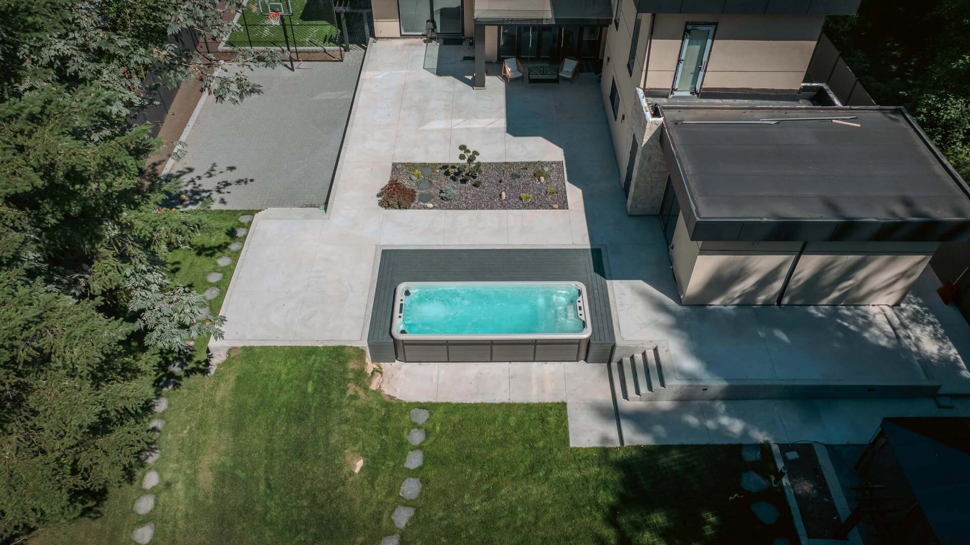 An aerial view of a modern backyard featuring a rectangular turquoise hot tub on a light gray concrete patio, adjacent to a green lawn and a contemporary house.
