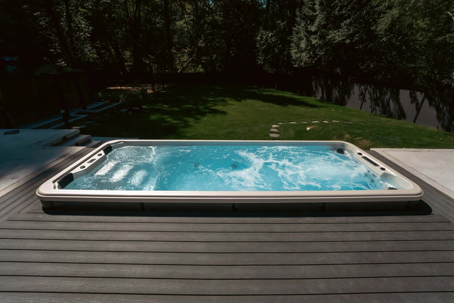 An outdoor hot tub with bubbling blue water sits on a dark gray composite deck in a backyard with green grass and trees in the background.