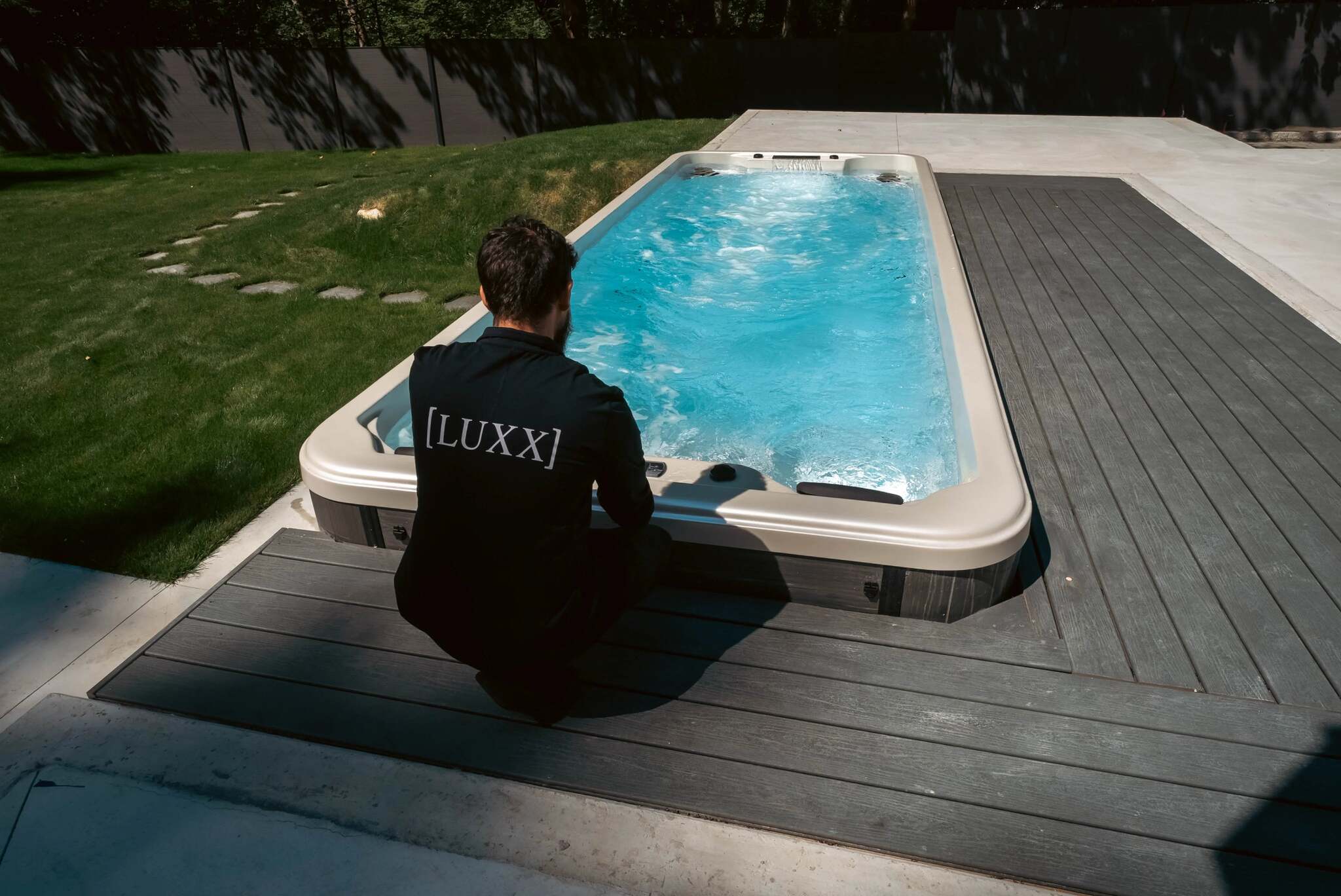 A man wearing a black t-shirt with '[LUXX]' printed on the back kneels beside a rectangular outdoor hot tub with blue water on a dark wood deck.