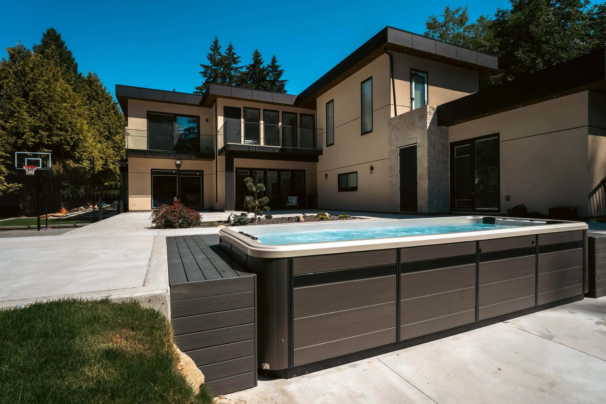 A modern outdoor hot tub with a dark gray wood-look exterior and light beige rim, situated on a concrete patio in front of a contemporary two-story house under a clear blue sky.