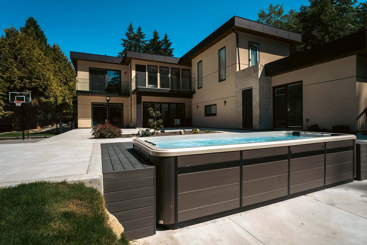 A modern outdoor hot tub with a dark gray wood-look exterior and light beige rim, situated on a concrete patio in front of a contemporary two-story house under a clear blue sky.