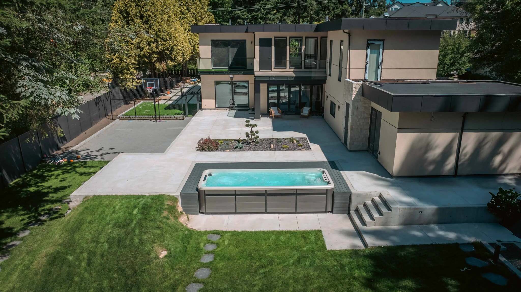 A modern two-story house with a large outdoor patio, a rectangular hot tub, a green lawn, and trees in the background.
